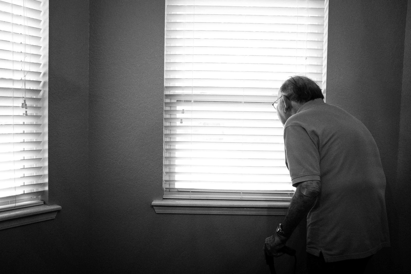 Adulto mayor mirando por la ventana de su casa, apoyado en un bastón